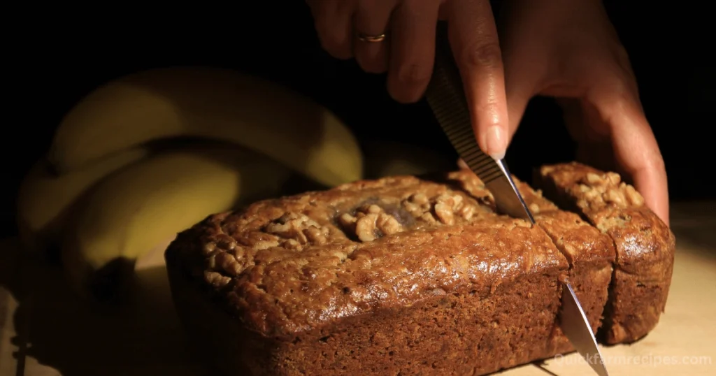 A freshly baked loaf of banana bread with a golden crust being sliced by hand under warm lighting, with ripe bananas in the background.