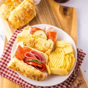 A plate of sliced herb-and-cheese bread sandwiches filled with deli meats, accompanied by chips.