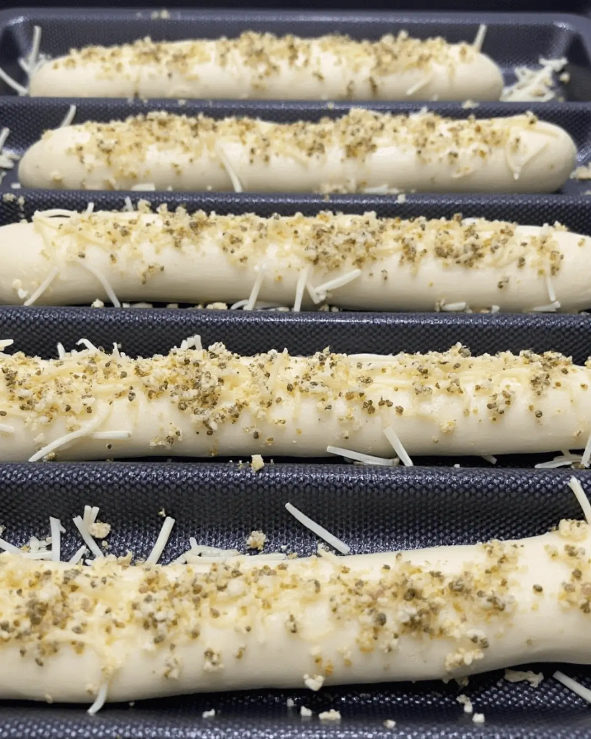 Rows of elongated bread dough on a perforated pan, sprinkled with shredded cheese and herbs before baking.